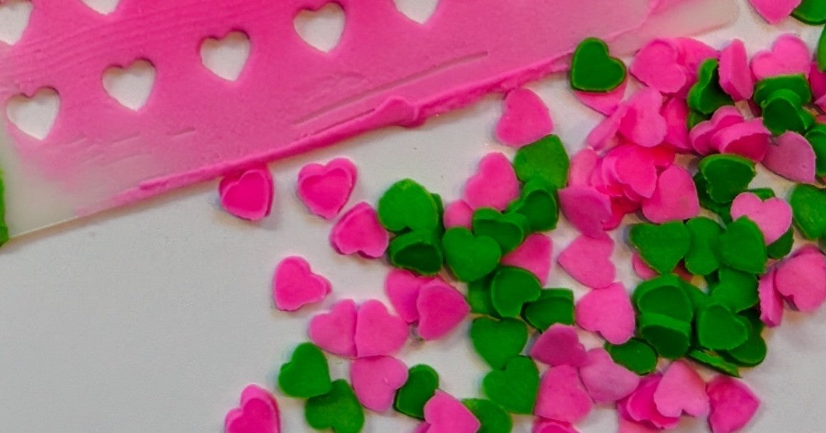 Pink and green homemade heart sprinkles beside a BakeArt Heart Sprinkle Stencil covered with royal icing