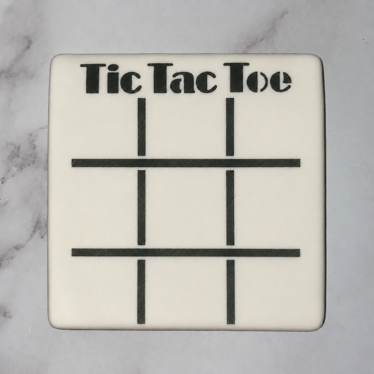 A square cookie, flooded in white icing and decorated with our Tic Tac Toe Game Cookie Stencil in black royal icing, BakeArt Stencils