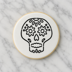  A white, round sugar cookie decorated with our Sugar Skull PYO Cookie Stencil in black royal icing and shown on a Carrara Marble countertop, BakeArt Stencils