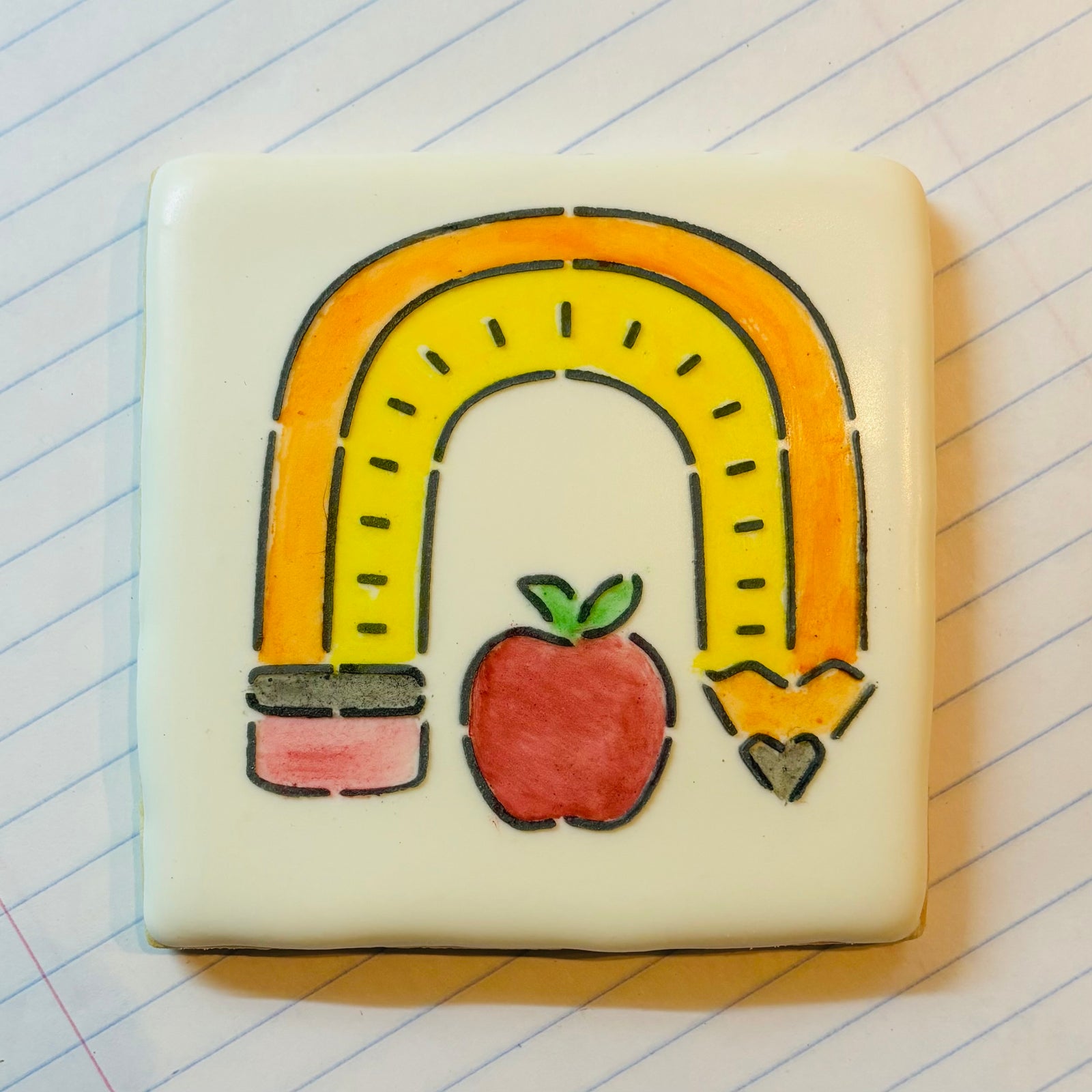 Pencil Rainbow PYO Stencil decorated on a white square cookie in black royal icing and then decorated with edible paint colors in assorted colors and shown on a sheet of notebook paper