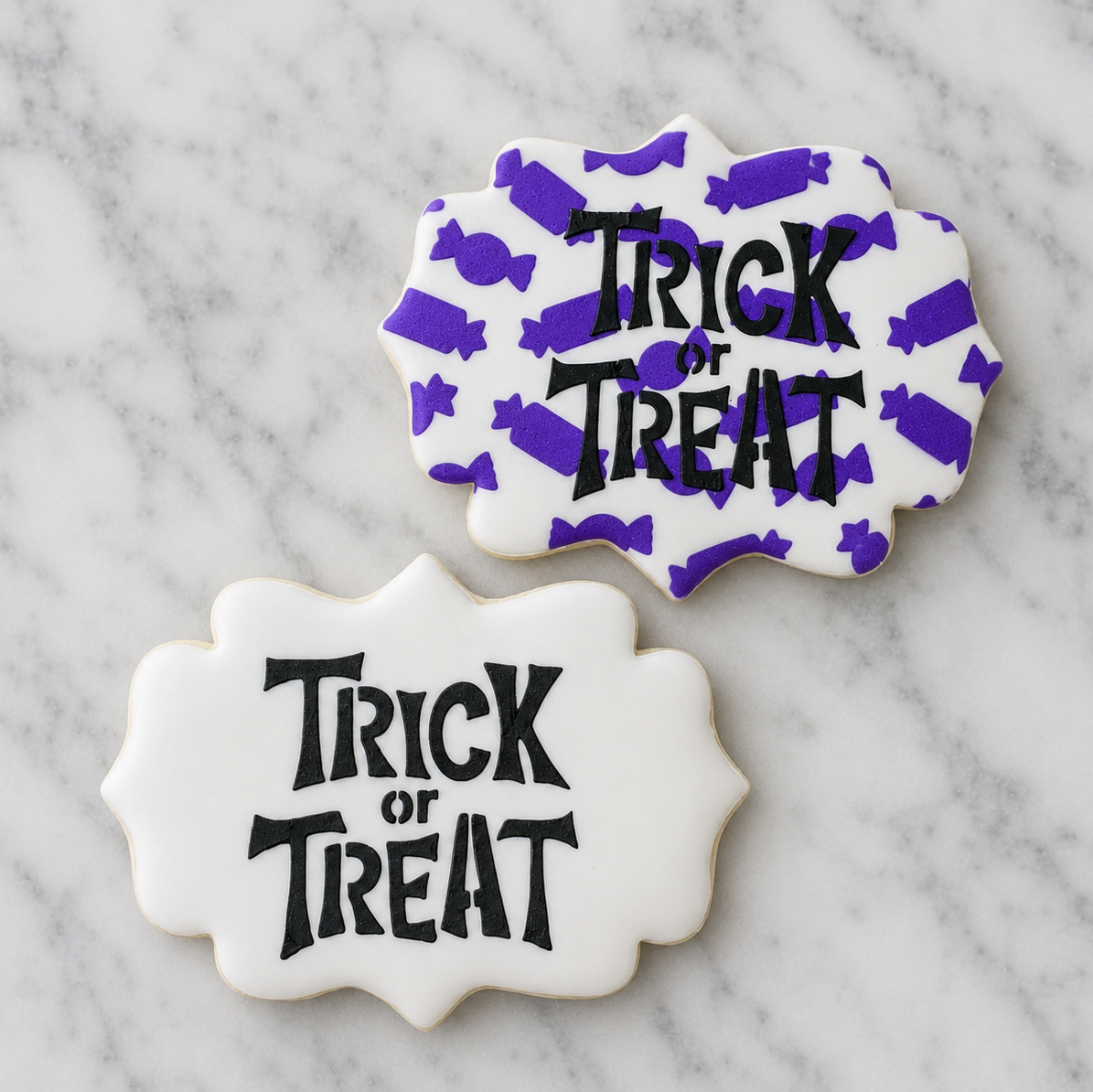 Two white, plaque shaped Halloween sugar cookies decorated with the Tricky Trick or Treat Cookie Stencil in black royal icing and one of the cookies also is stenciled with the Candy Pattern in purple. Both cookies shown on a Carrara Marble counter, BakeArt Stencils