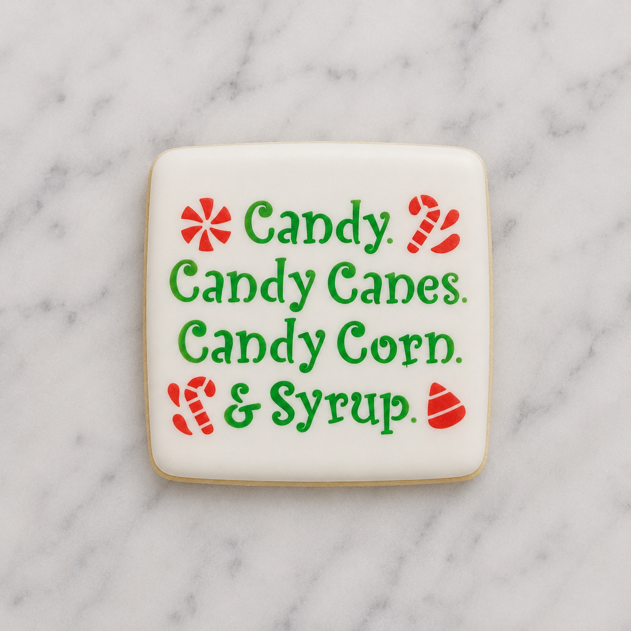 A white, square sugar cookie decorated with our Elf Candy Saying Cookie Stencil in green and red and lying on a marble counter