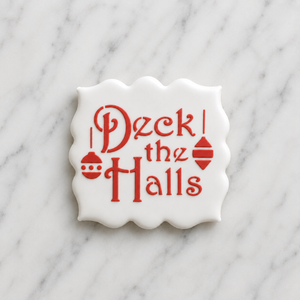 A square, plaque shaped cookie flooded in white icing and decorated with the Deck the Halls Cookie Stencil in red royal icing
