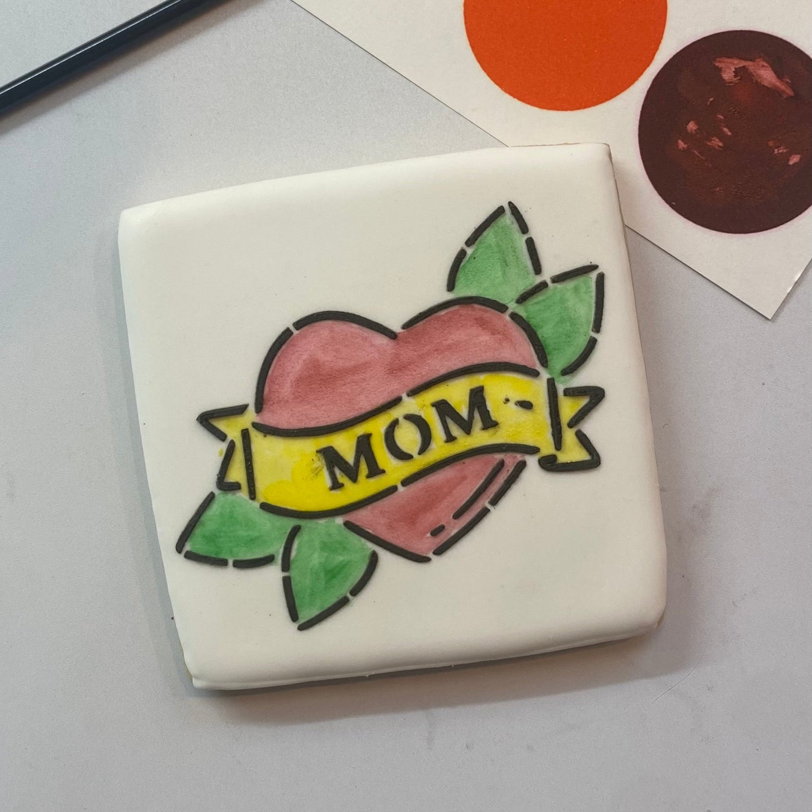 A square cookie flooded in white royal icing and decorated with our Mom Tattoo PYO Cookie Stencil in black royal icing and then painted in red, yellow and green edible food colors, BakeArt Stencils