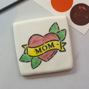 A square cookie flooded in white royal icing and decorated with our Mom Tattoo PYO Cookie Stencil in black royal icing and then painted in red, yellow and green edible food colors, BakeArt Stencils