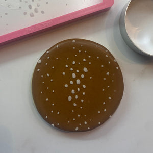 A round sugar cookie flooded in brown and stenciled with the Fawn Deer Spots Cookie Stencil in white royal icing and shown with the circle cookie cutter and decorating tools