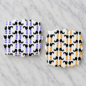 Two plaque shaped sugar cookies, flooded in white and airbrushed with the Witchy Witch Feet Cookie Stencil in purple, orange, and black and shown on a Carrara Marble counter, BakeArt Stencils