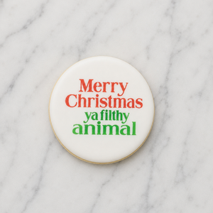 A white, round sugar cookie decorated with our Merry Christmas Filthy Animal Cookie Stencil in red and green and shown lying on a marble counter