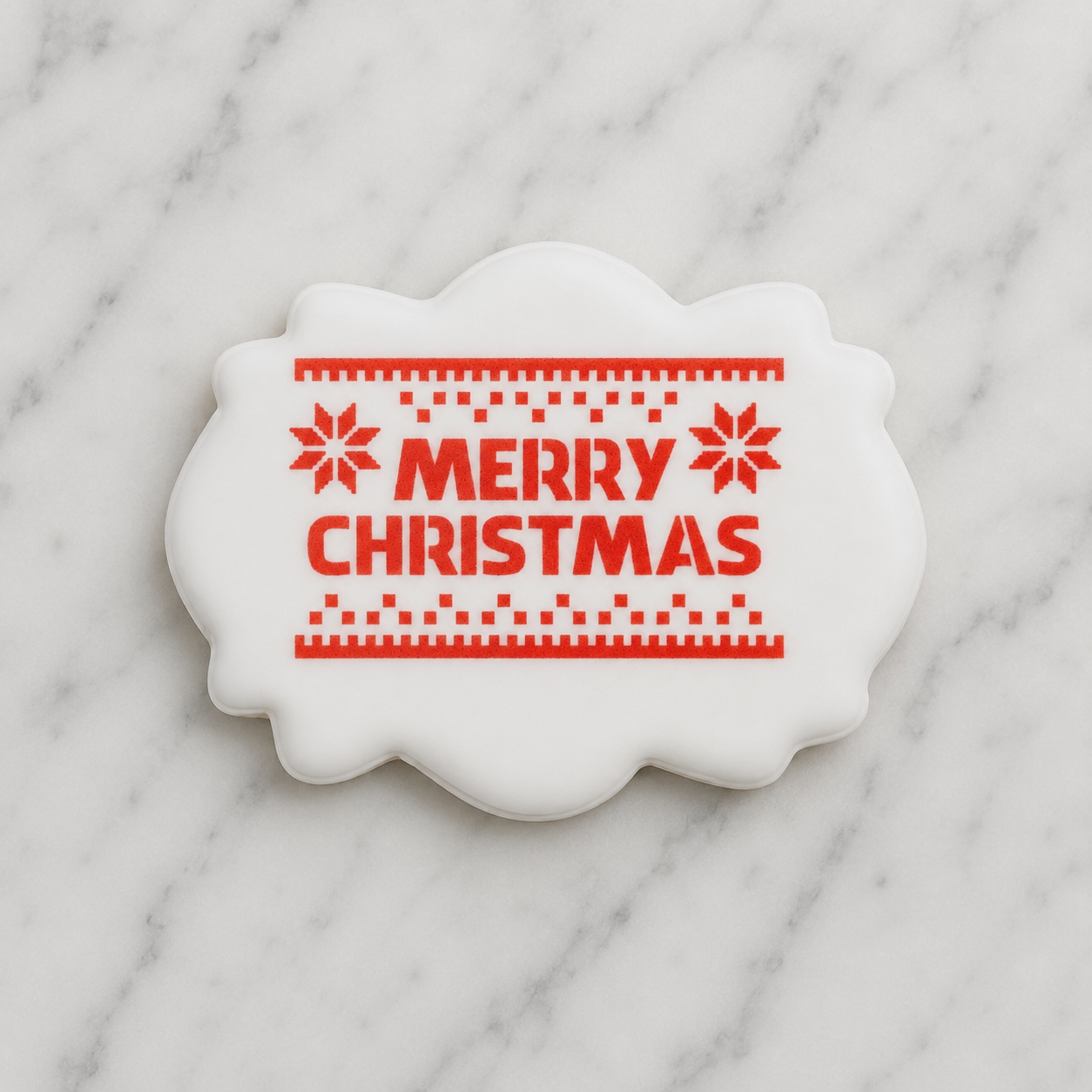 A white, plaque shaped cookie decorated with our Knitted Merry Christmas Cookie Stencil in red and shown on a marble counter