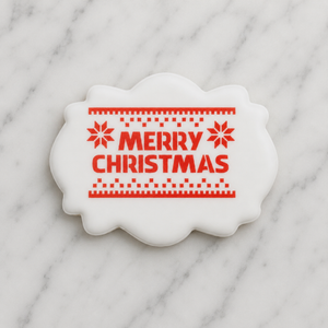 A white, plaque shaped cookie decorated with our Knitted Merry Christmas Cookie Stencil in red and shown on a marble counter