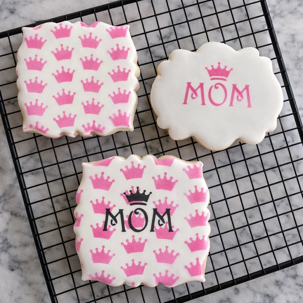 Three plaque shaped cookies all flooded in white, two are airbrushed with our Tiara Pattern Cookie Stencil in pink, one with our Queen Mom Cookie Stencil decorated in black royal icing and the plain cookie has the Queen Mom design in pink