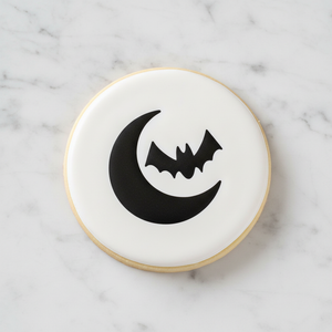 A white, round sugar cookie decorated with the Bat and Moon Cookie Stencil in black royal icing, laying on a marble counter, BakeArt Stencils