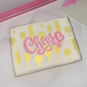 A rectangle shaped cookie flooded in white icing and airbrushed with our Bar Glasses Cookie Stencil in gold and then decorated with our Big Cheers Cookie Stencil in pink royal icing and sprayed with edible glitter
