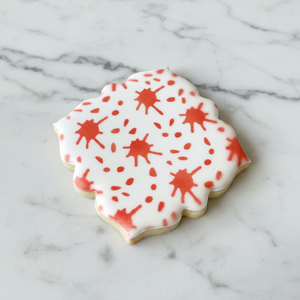 A white sugar cookie made with the Quinn Plaque Cookie Cutter and decorated with our Blood Splatter Cookie Stencil in red, BakeArt Stencils