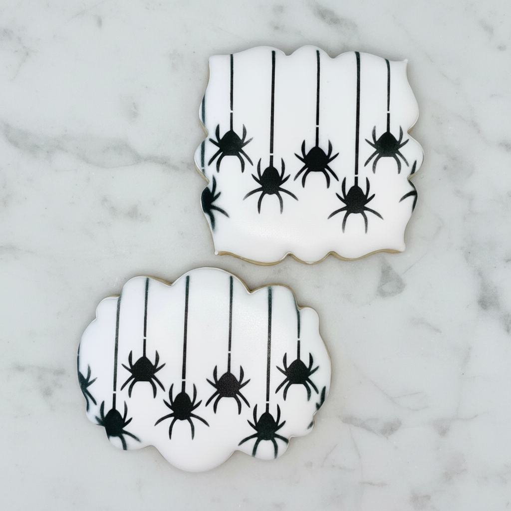 Twow white sugar cookies made with our plaque cookie cutters and decorated with the Spinning Spiders Cookie Stencil in black and laying on Carrara Marble, BakeArt Stencils