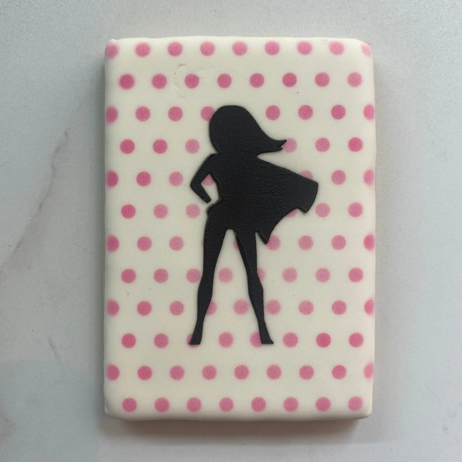 A rectangle shaped cookie flooded in white icing, airbrushed with our Teeny Dots Cookie Stencil and then stenciled with our Female Superhero Cookie Stencil in black royal icing.