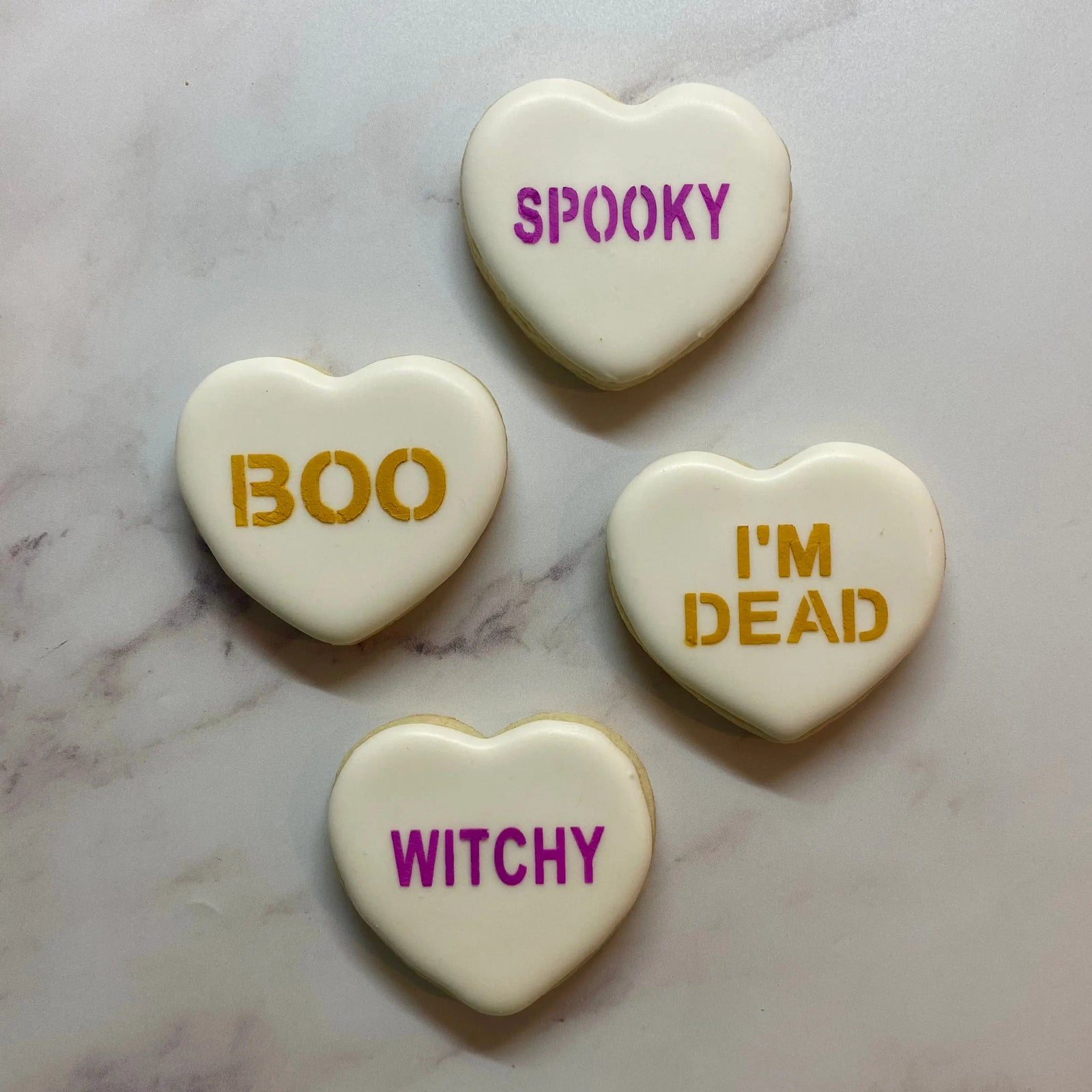 Four white heart-shaped sugar cookies decorated with the Halloween Conversation Sayings Cookie Stencil in orange and purple with the words spooky, boo, I'm dead and witchy and all laying on marble countertop, BakArt Stencils