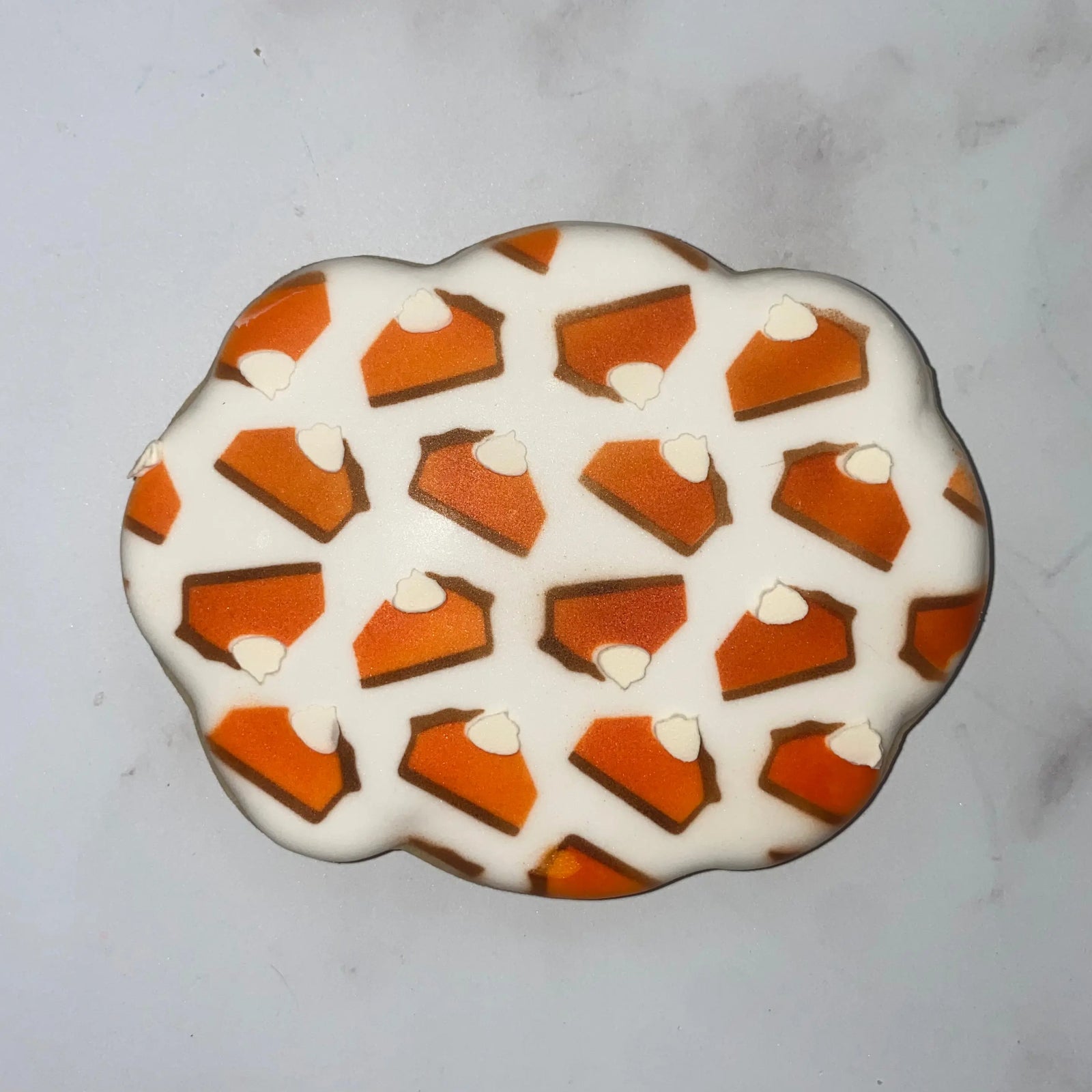 A plaque shaped cookie flooded in white icing and decorated with our Pie Slices Cookie Stencil Set decorated like pumpkin pie in shades of orange, brown and white, BakeArt Stencils
